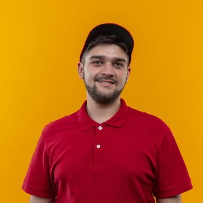 young delivery man in red uniform and cap looking at camera with smile on face standing over orange background